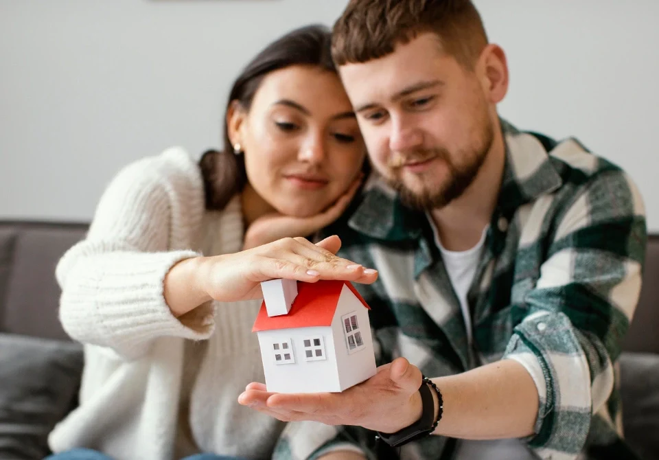Pareja joven protegiendo una maqueta de casa con las manos, simbolizando la seguridad que ofrece un seguro de impago de alquiler y sus requisitos principales.